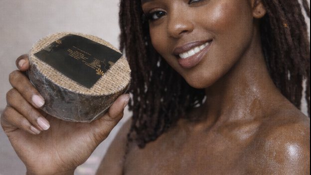 A woman with natural locs holding a bar of the client's own African black soap, warm studio light